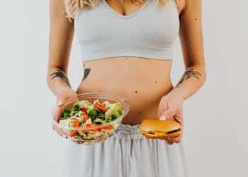 A woman in a sports bra holding a salad and cheeseburger, representing healthy vs junk food choices.