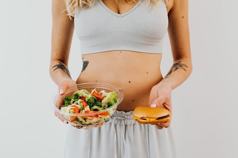 A woman in a sports bra holding a salad and cheeseburger, representing healthy vs junk food choices.