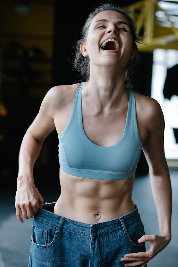 Fit young woman happily shows off weight-loss success by wearing oversized jeans in a gym.