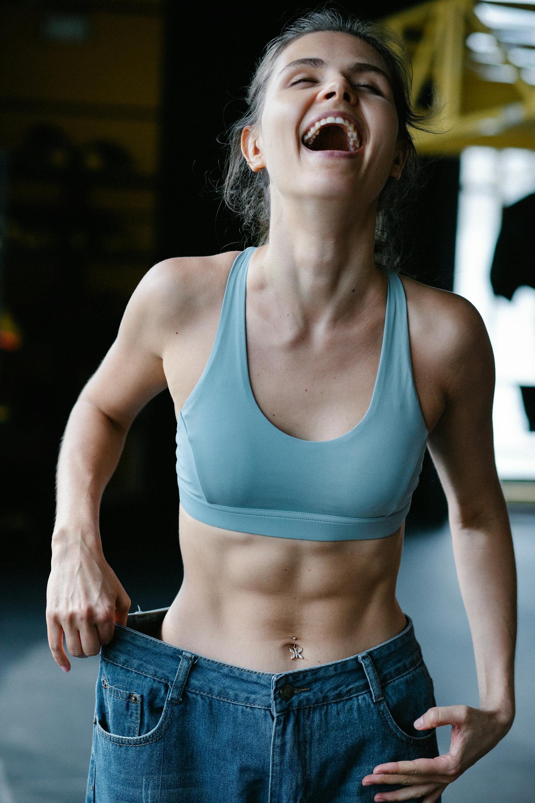 Fit young woman happily shows off weight-loss success by wearing oversized jeans in a gym.