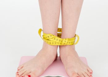 High angle of crop anonymous female with centimeter on legs checking weight on scales on white background