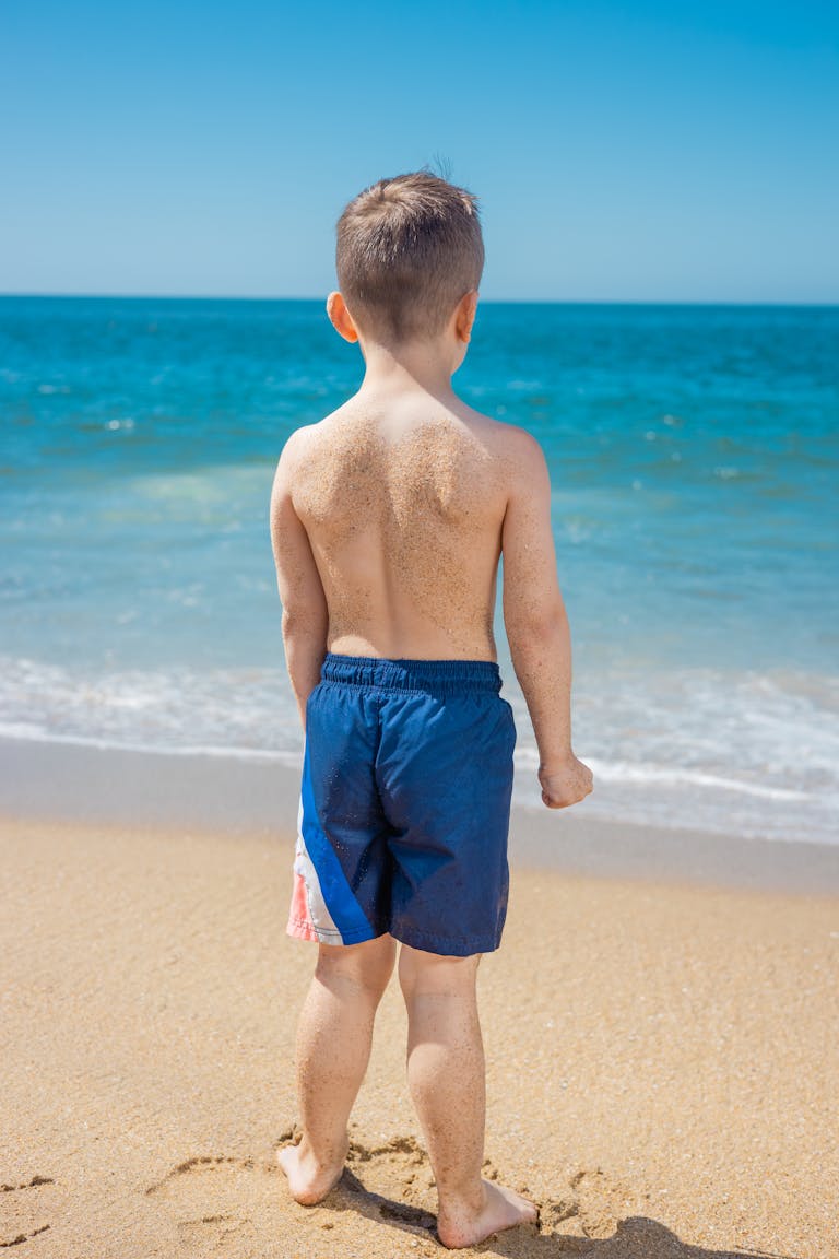 Young boy standing on sandy beach, gazing at the ocean on a sunny day.