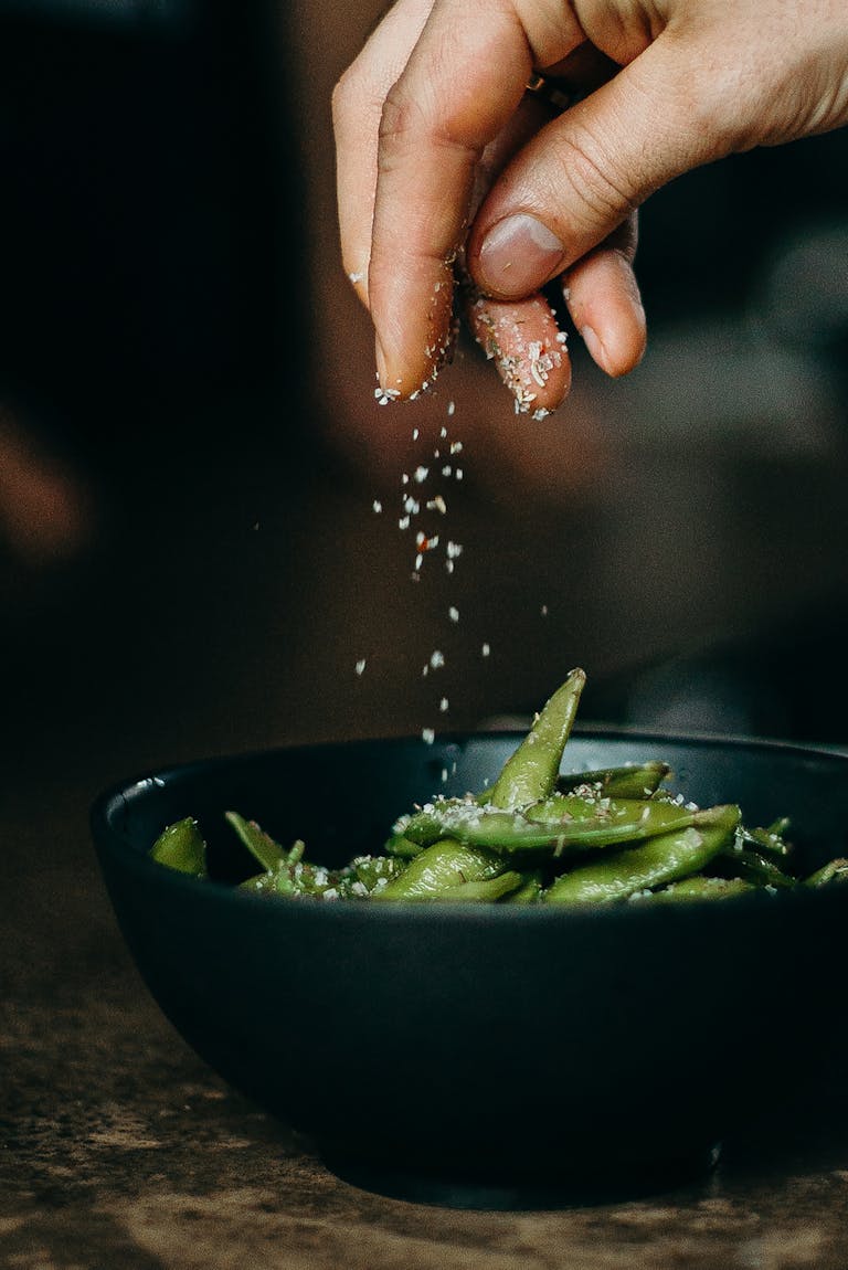 A hand sprinkles salt on fresh edamame, showcasing a delicious and healthy cooking moment.