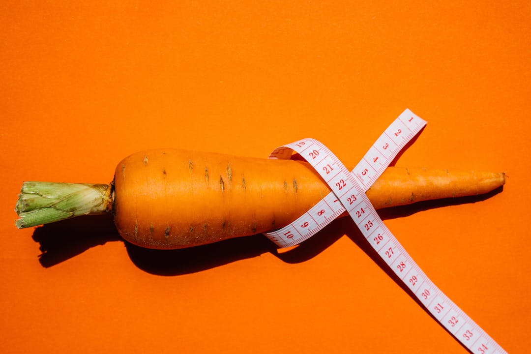 A vibrant image of a carrot wrapped with a measuring tape on an orange background, symbolizing health and nutrition.