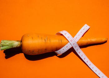 A vibrant image of a carrot wrapped with a measuring tape on an orange background, symbolizing health and nutrition.