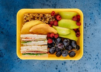Colorful lunch box with fruits, sandwiches, and a snack bar on a blue background.