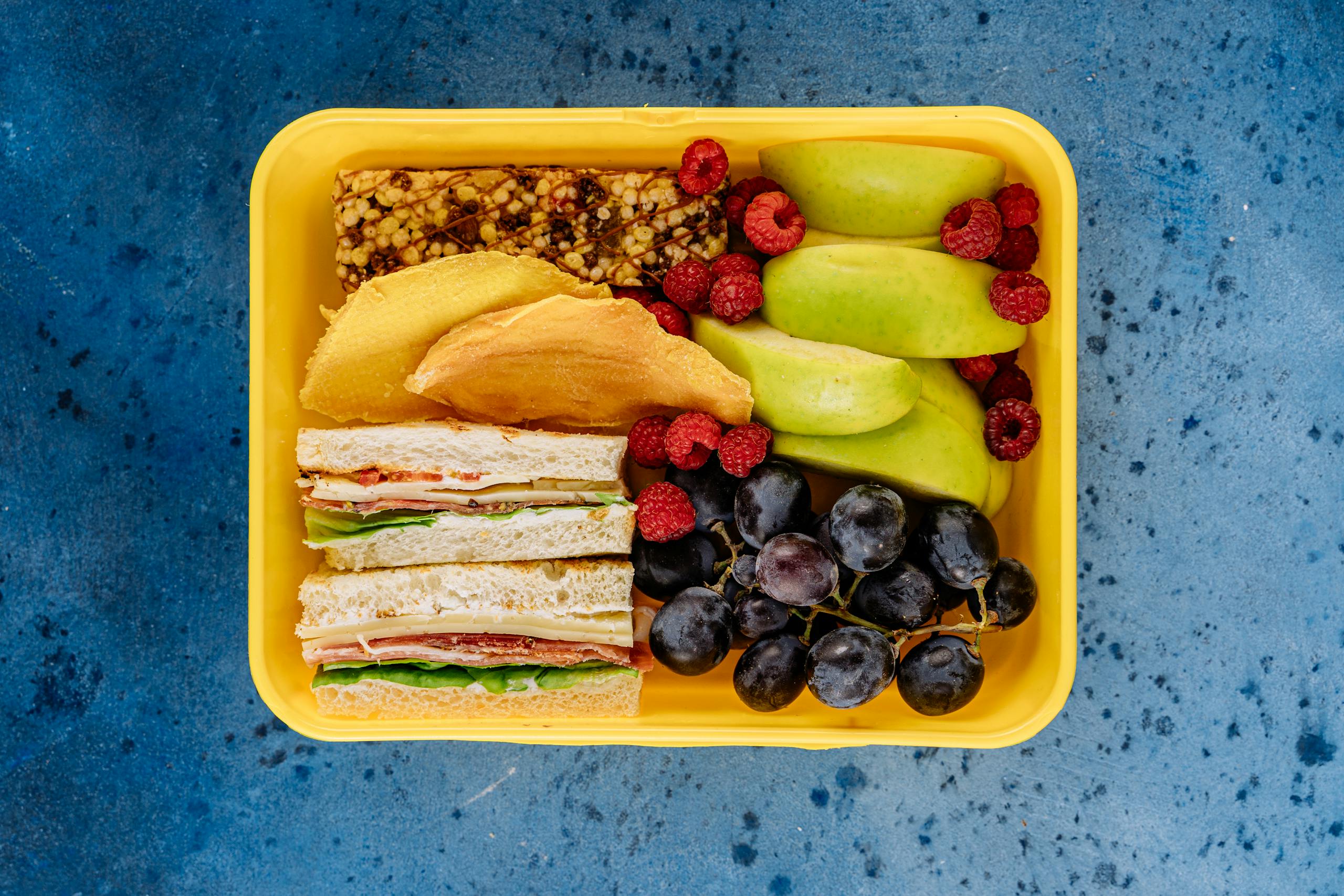 Colorful lunch box with fruits, sandwiches, and a snack bar on a blue background.